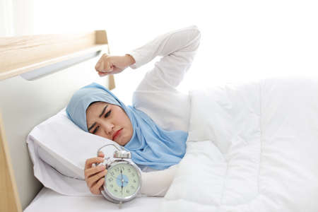Oversleeping asian muslim woman wearing white sleepwear lying on bed, missing ring of alarm clock waking up. side view portrait young girl trying to stop ringing while her head on pillowの写真素材
