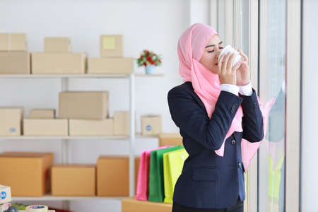 Religious asian muslim woman in blue suit drinking coffee, smiling and pink scarf on head with package box delivery background. Startup small business SME freelance girl work at home with happy faceの写真素材
