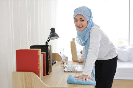 Indoor shot active young beautiful asian muslim housewife woman in sleepwear cleaning up wooden table with computer. Portrait cute girl with happy emotional cleaning bedroom in new apartment after moved in.の写真素材