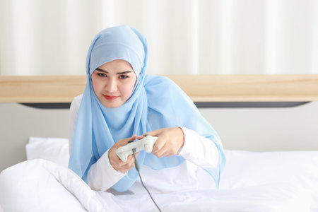 VDO game console station concept. active asian muslim woman wearing white sleepwear sitting on bed,...