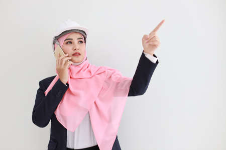 Smart muslim young asian woman wearing blue suit smiling confident, pointing and using mobile phone in studio. Isolated white background portrait with beautiful face girl. Advertisement concept.の写真素材