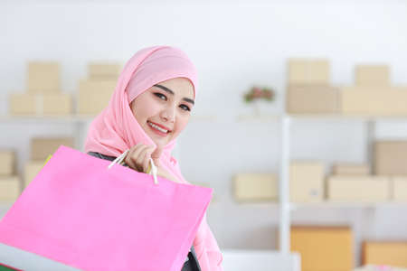 Active smart asian muslim woman in jean jacket standing and holding shopping bags with online package box delivery background. Beautiful girl looking at camera and smile. Shopping conceptの写真素材