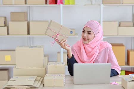 Active asian muslim woman in blue suit sitting and working with computer and showing online package box delivery with happy smiling face. Startup small business freelance working girl look at product.の写真素材