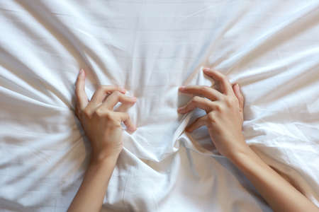 Top view young sexy woman hands pulling white sheets in ecstasy in hotel. Cute girl doing sign orgasm on white bed, sex and erotic concept for advertisementの写真素材