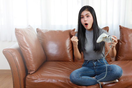 VDO game console station concept. active asian woman sitting on sofa, holding joystick and playing...