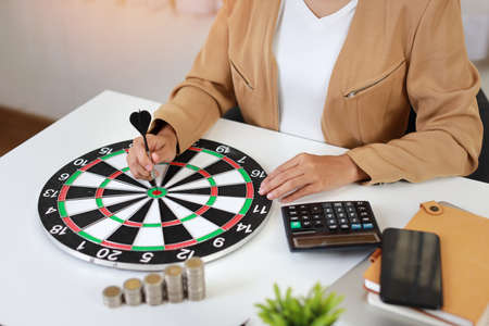 Smart and active asian businesswoman hands putting dart to target dartboard with saving growth on stacking coin on table, meaning of earning money or investment or tax for business advertising conceptの写真素材