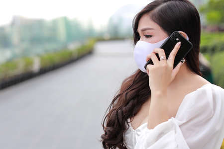Young asian business woman in white casual dress with protect mask for healthcare, walking in public outdoor and working on smartphone. New normal and social distancing conceptの写真素材