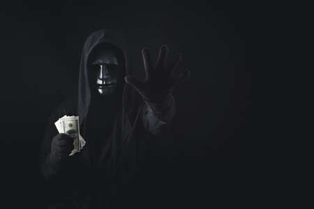 Dangerous anonymous hacker man in hooded showing stop gesture and holding banknote, break security data and hack password with personal Bank account. Internet crime cyber attack security conceptの写真素材