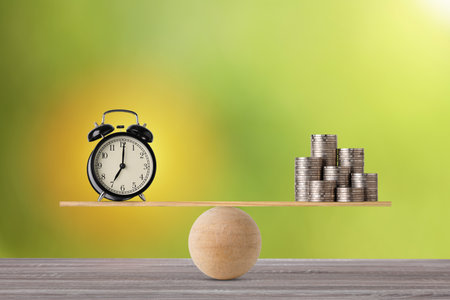 Alarm clock on seesaw balancing with stacking coins money on wooden table, meaning of business investment and saving growth with time for advertising concept.の写真素材