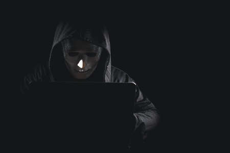 Dangerous anonymous hacker man in black hooded, white mask use computer and mobile phone, break security data corporate server. He working on black background. Internet crime, cyber attack security.の写真素材