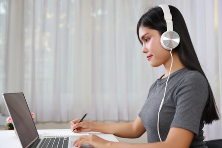 Young smart and active asian female student sitting at table with headset, using computer and taking note for studying online with happy face (new normal concept)の写真素材