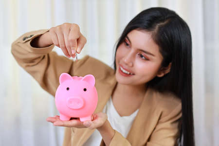 Smart and active asian businesswoman putting coin into piggy bank (focus on hand), meaning of earning money or investment or tax for business advertising conceptの写真素材