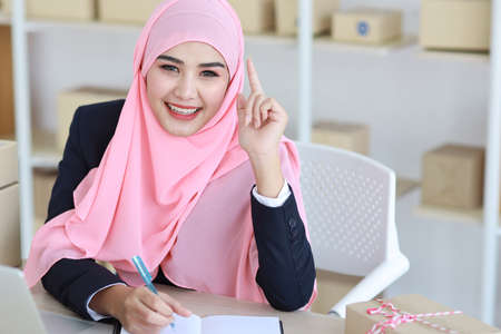 Active asian muslim woman in blue suit sitting and working with computer and online package box delivery. Startup small business freelance working girl look at camera with smilingの写真素材