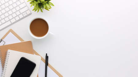 Top view white wooden workspace office desk with computer and office supplies. Flat lay work table with blank notebook, keyboard, green leaf and coffee cup. Copy space for your advertising content.の写真素材