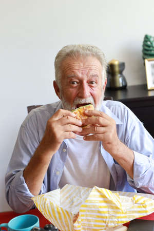 full length of happy caucasian elderly sitting and eating hamburger in living room with smiling faceの写真素材
