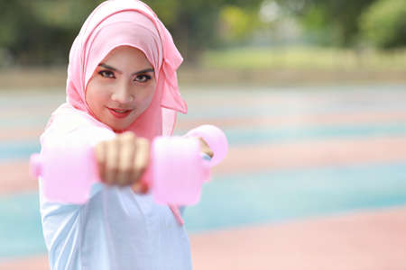 Athletic young asian muslim woman in sportswear standing and lifting dumbbells outdoor for morning exercise. Active girl work out on biceps exercise with green tree background. Sport conceptの写真素材