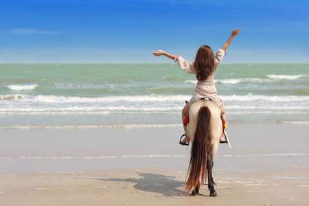 Rear view of young asian beautiful on horse standing on the sandy beach in brown dress. Cute girl raising hand and having fun on holiday vacation with happy time. Free space for advertising.の写真素材