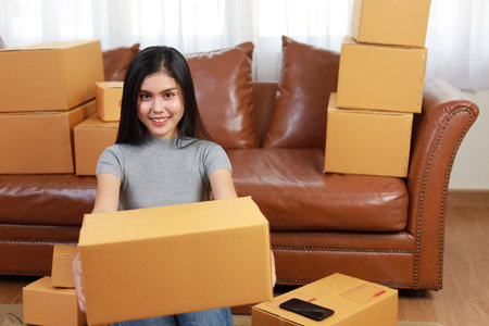 Young asian smart and active woman in casual dress holding box packaging and working from home with smartphone and online purchase shopping order (new normal concept)の写真素材