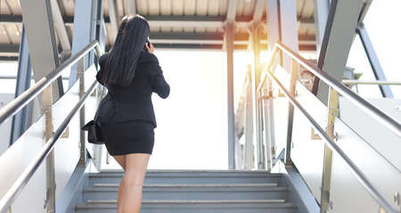 Rear view asian business woman in business black suit with protect mask for healthcare walking on footbridge public outdoor and using smartphone. New normal and social distancing conceptの写真素材