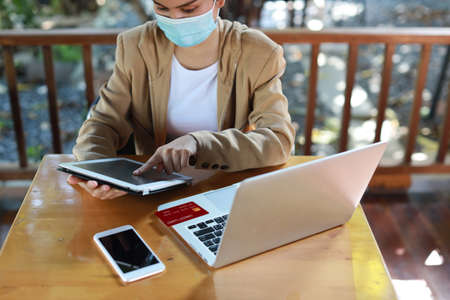 Young asian woman hands in casual dress with protect mask for healthcare, sitting in coffee shop and using tablet and working on laptop computer. New normal and social distancing conceptの写真素材