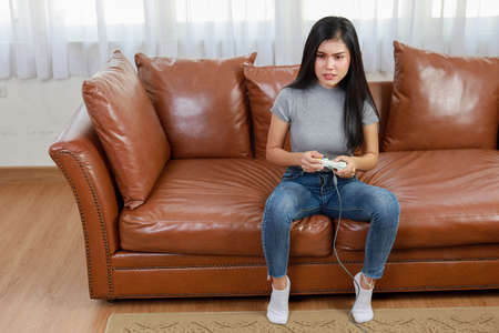 VDO game console station concept. active asian woman sitting on sofa, holding joystick and playing exciting game. Cute girl looked excited with game controller consoleの写真素材