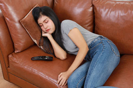 TV and happiness concept. Beautiful asian woman in casual lying on sofa in living room, holding television remote and sleeping while watching television with happy smiling face. Lifestyle concept.の写真素材