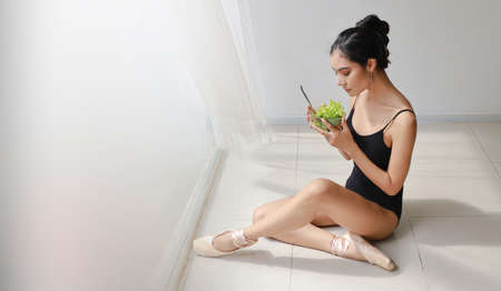 beautiful healthy and sporty asian young woman holding salad bowl and eat after training ballet dancing while sitting with relaxationの写真素材