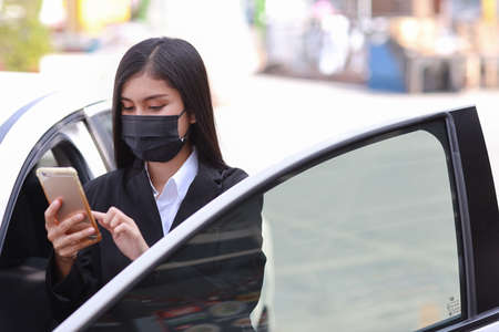 Young asian healthy woman in business black suit with protect mask for healthcare get off automobile and using smartphone. New normal and social distancing conceptの写真素材