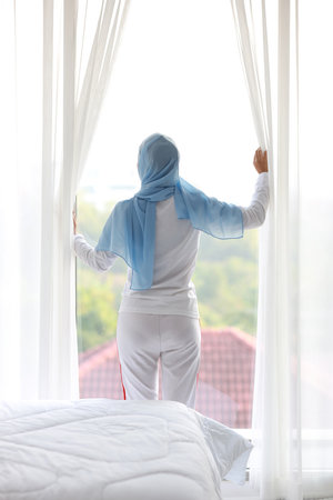 Rear view beautiful asian woman wearing white muslim sleepwear, stretching her arms after getting up in the morning at sunrise. Cute young girl with blue hijab standing and relaxing while looking awayの写真素材
