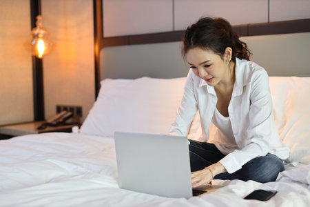 Active asian woman in casual sitting on bed and working on computer with notebook in hotel, beauty businesswoman with bedroom background. Lifestyle technology conceptの写真素材