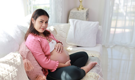 Happy smiling young asian pregnant woman sitting and resting on sofa in living room while touching her belly and looking camera. Expectant mother preparing and waiting for baby birth during pregnancy.の写真素材