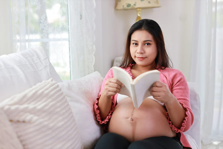 Happy young asian pregnant woman sitting and resting on sofa in living room while reading book and...