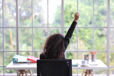 Rear view happy business woman raising hand with office accessories on table. Curly hair and successful businesswoman sitting and working on computer with exciting emotion at office. Success.の写真素材