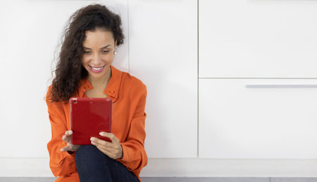 Happy smiling latin woman sitting and using internet on tablet in living room. Lifestyle with technologyの写真素材