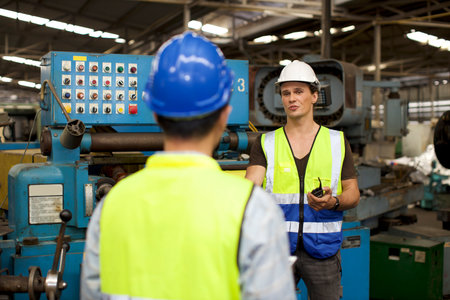 Technician engineer or worker man in protective suit standing and using walkie talkie radio while controlling or maintenance operation work lathe metal machine at heavy industry manufacturing factoryの写真素材