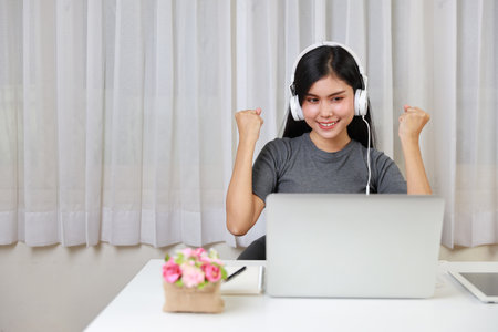 Young smart and active asian female student sitting at table with headset, using computer and taking note for studying online with exciting emotion (study online concept)の写真素材