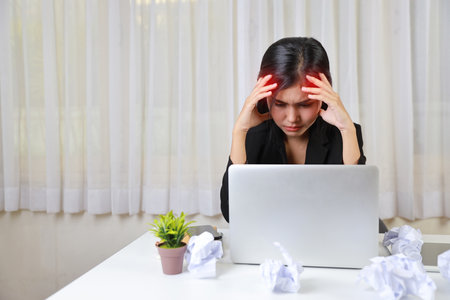 Mid adult businesswoman touching head on table after bad news business failure or get fired and feeling discouraged, distraught and hopeless on blue background with computer and crumpled paperの写真素材