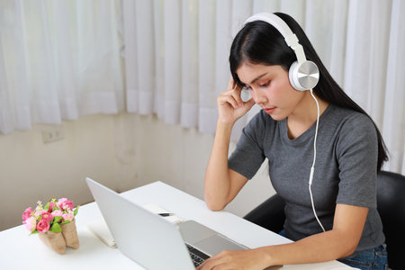 Young smart and active asian female student sitting at table with headset, using computer and taking note for studying online with serious face (study only concept)の写真素材