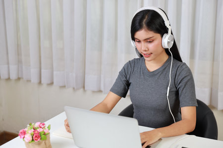 Young smart and active asian female student sitting at table with headset, using computer and taking note for studying online with happy smiling face (study only concept)の写真素材