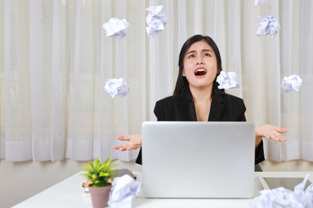 Mid adult businesswoman screeming on table after bad news business failure or get fired and feeling discouraged, distraught and hopeless on blue background with computer and falling crumpled papersの写真素材