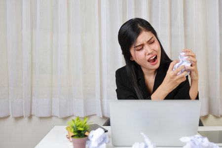 Mid adult businesswoman screeming on table after bad news business failure or get fired and feeling discouraged, distraught and hopeless on blue background with computer and crumpled paperの写真素材