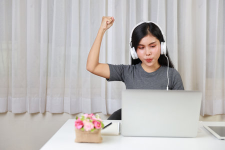 Young smart and active asian female student sitting at table with headset, using computer and taking note for studying online with exciting emotion (study online concept)の写真素材