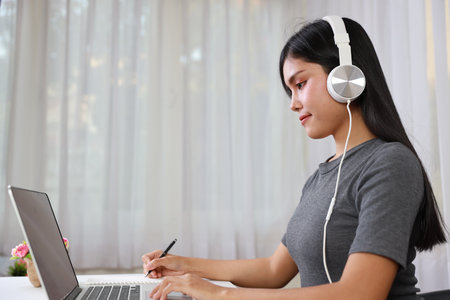 Young smart and active asian female student sitting at table with headset, using computer and taking note for studying online with happy face (study online concept)の写真素材