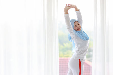 Beautiful asian woman wearing white muslim sleepwear, stretching her arms after getting up in the morning at sunrise. Cute young girl with blue hijab standing and relaxing with happy and smiling face.の写真素材
