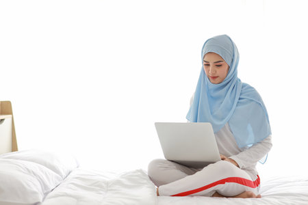 Young asian woman use laptop sitting on bed in bedroom. Beautiful girl in white muslim sleepwear with blue hijab check order or study from notebook before go to bed. Lifestyle and technology conceptの写真素材