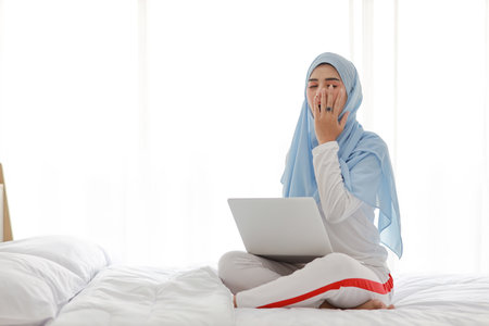 Young asian woman using computer sitting on bed in bedroom. Beautiful girl in muslim sleepwear yawning after working before go to bed. Lifestyle and technology conceptの写真素材