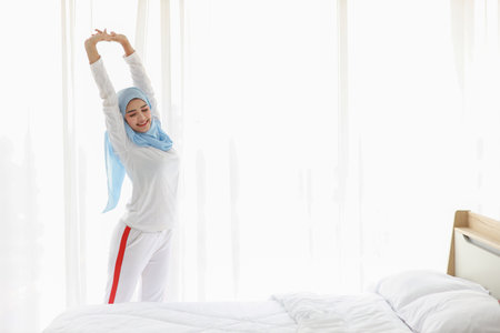 Beautiful asian woman wearing white muslim sleepwear, stretching her arms after getting up in the morning at sunrise. Cute young girl with blue hijab standing and relaxing with happy and smiling face.の写真素材