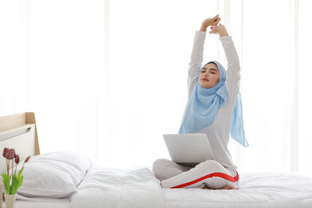 Young asian woman using computer sitting on bed in bedroom. Beautiful girl in muslim sleepwear with blue hijab stretching her arms after working before go to bed. Lifestyle and technology conceptの写真素材