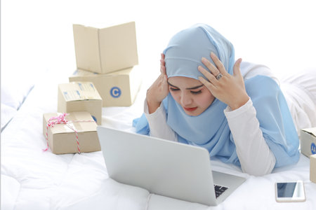 Beautiful and young asian woman in muslim sleepwear with headache emotion lies on bed with computer, mobile phone and online package box delivery. Smart girl with hijab receive bad news and tiredの写真素材