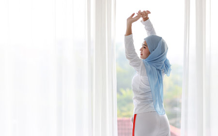Beautiful asian woman wearing white muslim sleepwear, stretching her arms after getting up in the morning at sunrise. Cute young girl with blue hijab standing and relaxing with happy and smiling face.の写真素材